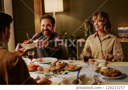 Biracial company of friends sitting at table with different treats and dishes eating them, cheerful an offering plate with baked potatoes to his friend Biracial company of friends sitting at table with different treats and dishes eating them, cheerful an offering plate with baked potatoes to his friend 120215257