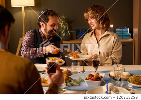 Jewish husband putting delicious food on wifes plate while they and their friends celebrating holiday together Jewish husband putting delicious food on wifes plate while they and their friends celebrating holiday together 120215259