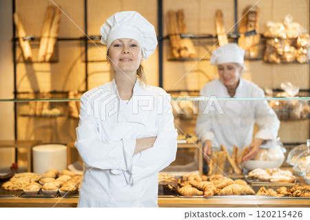 Smiling female bakery worker in uniform standing in front of pastry stand in bakeshop Smiling female bakery worker in uniform standing in front of pastry stand in bakeshop 120215426