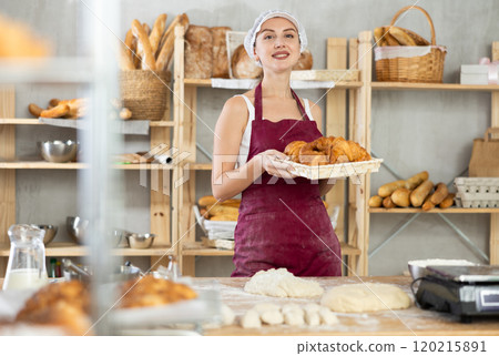 Smiling female baker in white uniform stands behind counter in small private bakery offering croissants 120215891