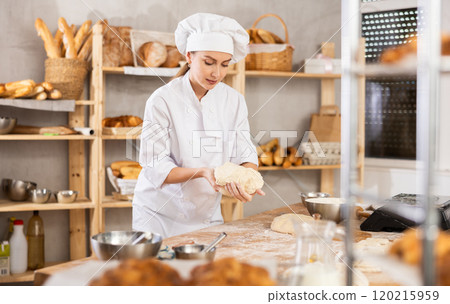 Skilled smiling young girl professional baker in white uniform standing at work table and kneading dough during working day in bakery 120215959
