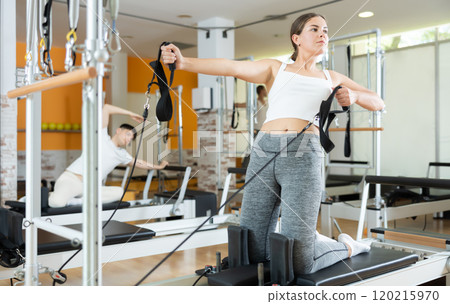 Girl in gym with help of rope reformer performs exercises to strengthen muscles of her arms. 120215970