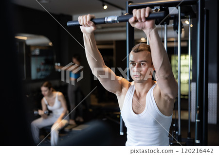 Slender athletic man work out muscles of arms and shoulders on simulator in gym of fitness club Slender athletic man work out muscles of arms and shoulders on simulator in gym of fitness club 120216442