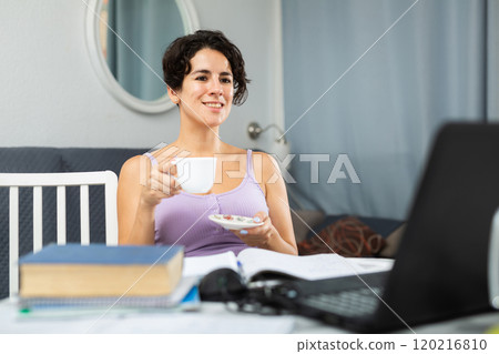 Laughing satisfied woman with cup of coffee in front of laptop at table 120216810