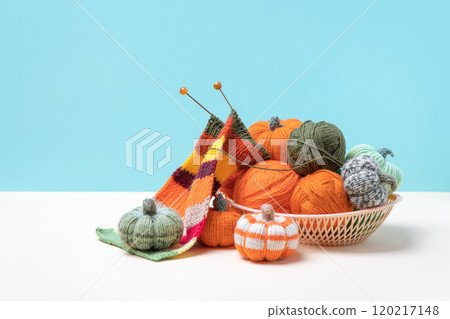 Green and orange knitting balls, orange pumpkins, striped scarf in a basket with knitting needles on beige background. Set for the hobby of knitting warm clothes or toys. Concept needlework 120217148