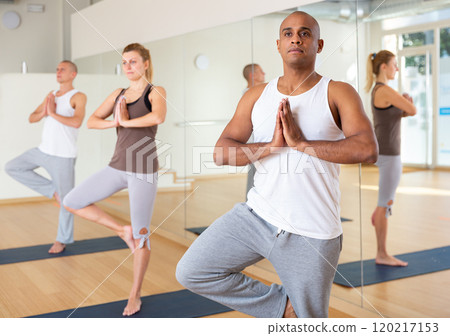 Hispanic man standing in Eka Pada Pranamasana during group yoga training Hispanic man standing in Eka Pada Pranamasana during group yoga training 120217153