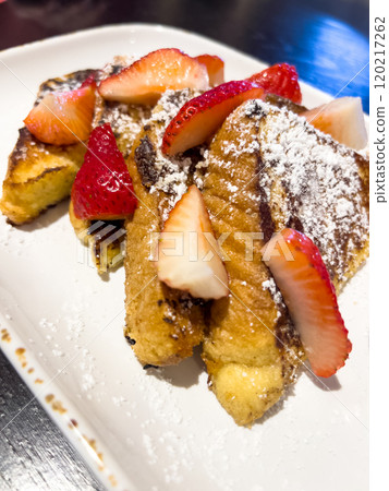 A delicious serving of French toast topped with fresh strawberries and a dusting of powdered sugar, served on a rectangular plate with a small pitcher of syrup on the side. The crispy, golden-brown A delicious serving of French toast topped with fresh strawberries and a dusting of powdered sugar, served on a rectangular plate with a small pitcher of syrup on the side. The crispy, golden-brown 120217262