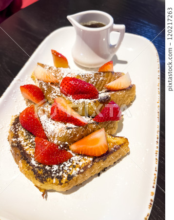 A delicious serving of French toast topped with fresh strawberries and a dusting of powdered sugar, served on a rectangular plate with a small pitcher of syrup on the side. The crispy, golden-brown A delicious serving of French toast topped with fresh strawberries and a dusting of powdered sugar, served on a rectangular plate with a small pitcher of syrup on the side. The crispy, golden-brown 120217263
