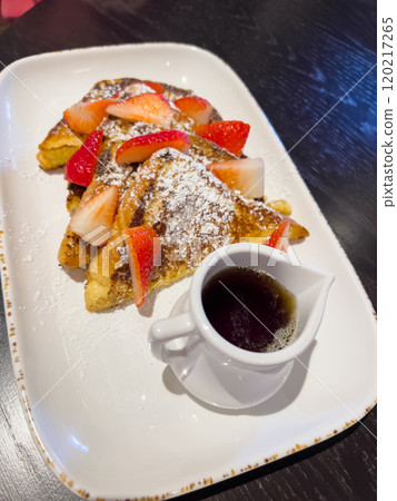 A delicious serving of French toast topped with fresh strawberries and a dusting of powdered sugar, served on a rectangular plate with a small pitcher of syrup on the side. The crispy, golden-brown 120217265