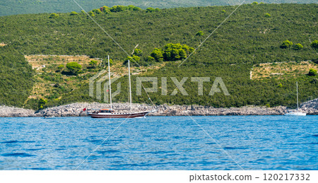 A peaceful sailing boat anchored near a lush green coastline with hills and blue waters. 120217332
