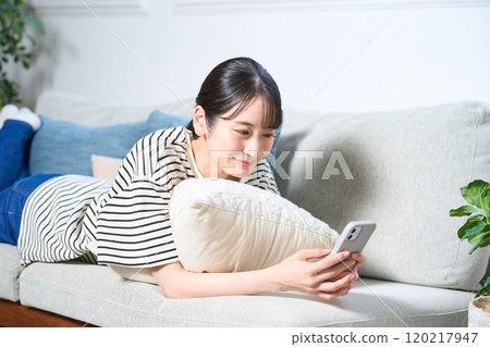 A young woman in casual clothes lying on a sofa, living room, smartphone 120217947