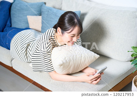 A young woman in casual clothes lying on a sofa, living room, smartphone 120217953
