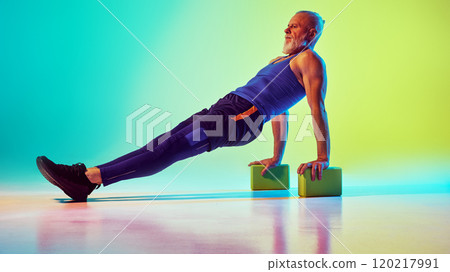 Sportive senior man in sportswear training, doing exercised with yoga blocks against gradient green yellow background in neon light 120217991