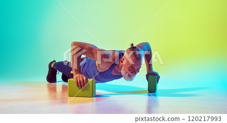 Athletic senior man in sportswear training, doing push ups yoga blocks against gradient green yellow background in neon light 120217993