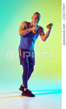 Kickboxing. Full-length of elderly sportive man training with dumbbell against gradient green yellow background in neon light 120217997