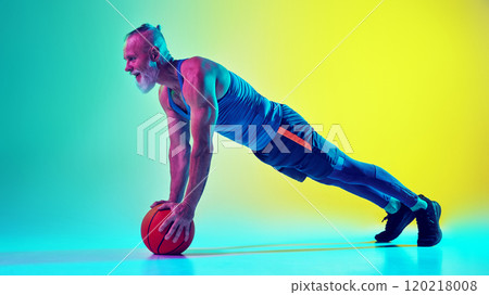 Muscular elderly man in sportswear standing on plank pose on basketball ball against gradient green yellow background in neon light. Side view 120218008
