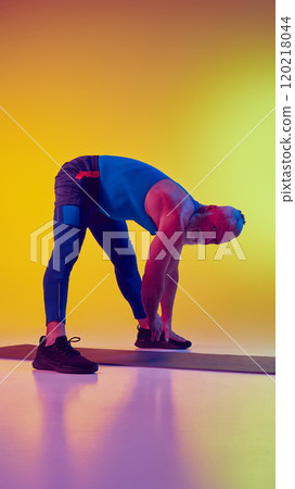 Vertical image of muscular athletic senior man in sportswear, standing on fitness mat and training against gradient yellow background in neon light Vertical image of muscular athletic senior man in sportswear, standing on fitness mat and training against gradient yellow background in neon light 120218044