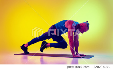 Muscular elderly man in sportswear training on fitness mat, doing mountain climber exercises against gradient yellow background in neon light 120218079