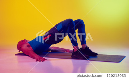 Senor man in sportswear lying on fitness mat and doing glute bridge exercise for fit lower body against gradient yellow background in neon light 120218083