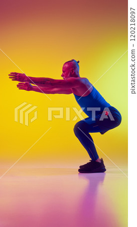Side view full-length image of elderly man in sportswear training, doing squats exercises against gradient yellow background in neon light Side view full-length image of elderly man in sportswear training, doing squats exercises against gradient yellow background in neon light 120218097