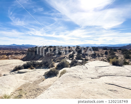 A sweeping view from the San Bench View Area reveals a vast desert landscape with rugged rock formations, scattered vegetation, and a distant mountain range under a clear blue sky. The rocky terrain 120218749