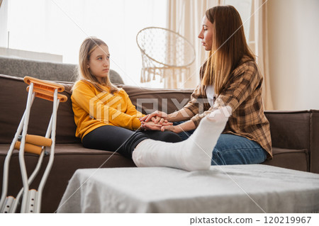 Girl with broken leg in cast sits with mother on couch at home. Sad little kid hugging teddy bear recovering from broken leg. Girl using cast crutches for quick rehabilitation. Mom supporting daughter 120219967