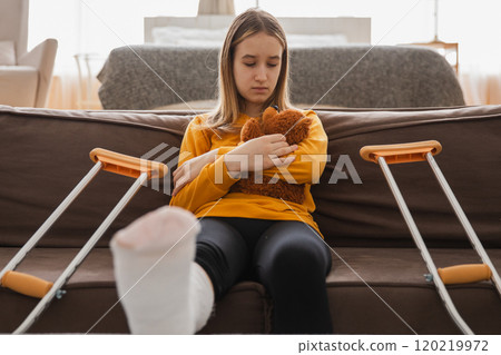 Young girl with broken leg cast sitting on sofa. Sad little kid hugging her teddy bear recovering from broken leg at home. Girl using cast and crutches. Accident injury fracture treatment foot bandage 120219972