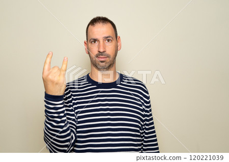 Portrait of bearded man showing rock and roll gesture, heavy metal sign, exclaiming with joy. Indoor studio photo isolated on beige background. 120221039