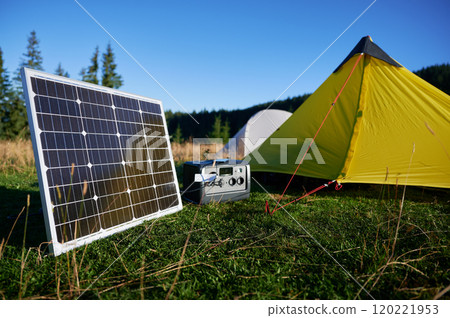 Tourist tent with solar panel and portable power station nearby in grassy field. Scene surrounded by rolling hills, tall pine trees under clear blue sky, showcasing eco-friendly camping setup. 120221953