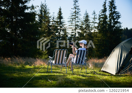 Woman camper relaxes next to tent with photo camera. Female tourist surrounded by tall evergreen trees and bathed in soft evening light, capturing peacefulness of moment. 120221954