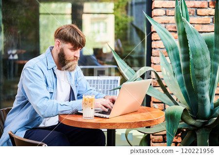 Man works on laptop at outdoor round table, with glass of iced coffee. Solar panel nearby, emphasizing sustainable, modern eco-friendly workspace that combines technology and sustainability. 120221956