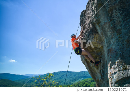 Aerial view of male rock climber ascending rugged limestone cliff with harness and rope for safety. Sportsman climbing on vertical large boulder at Dobvush Rocks in Carpathian mountains, Ukraine. 120221971