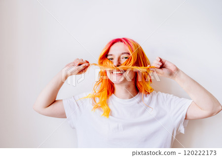 Cheerful young adult woman play with bright red and yellow hair against white background wearing plain white shirt, smiling playfully. Happiness and fun, creating lively, carefree atmosphere concept. Cheerful young adult woman play with bright red and yellow hair against white background wearing plain white shirt, smiling playfully. Happiness and fun, creating lively, carefree atmosphere concept. 120222173