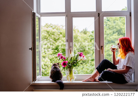 Young woman with bright hair sitting by large window, sipping coffee while enjoying tranquil view. Cat sitting nearby with vase of flowers, peaceful moments. Young woman with bright hair sitting by large window, sipping coffee while enjoying tranquil view. Cat sitting nearby with vase of flowers, peaceful moments. 120222177