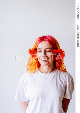 Portrait of smiling young Caucasian woman with vibrant pink and orange hair in white t-shirt, bright orange flowers behind her ears. Happiness, creativity, youthful energy, against clean background. Portrait of smiling young Caucasian woman with vibrant pink and orange hair in white t-shirt, bright orange flowers behind her ears. Happiness, creativity, youthful energy, against clean background. 120222178