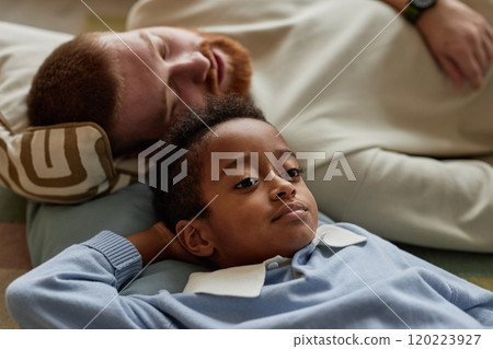 Portrait shot of thoughtful little Black boy resting with adoptive father lying down on floor in living room while enjoying time together on weekend, copy space Portrait shot of thoughtful little Black boy resting with adoptive father lying down on floor in living room while enjoying time together on weekend, copy space 120223927