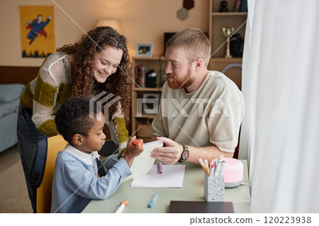 Medium shot of happy adoptive mother and father engaged in hand drawing with little Black boy sitting at desk in kids room Medium shot of happy adoptive mother and father engaged in hand drawing with little Black boy sitting at desk in kids room 120223938