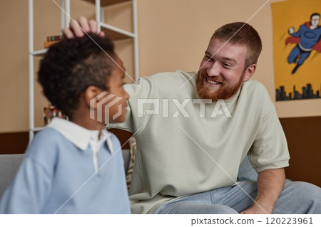Medium shot of cheerful dad looking with adoring smile at little adopted kid praising son while sitting on sofa in boys room 120223961