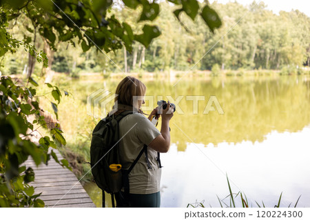 A young female photographer uses a camera to capture a serene lake scene. Travel and hobby concept. Woman with backpack is photographing a landscape near to river A young female photographer uses a camera to capture a serene lake scene. Travel and hobby concept. Woman with backpack is photographing a landscape near to river 120224300