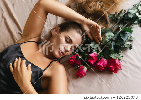 A woman is laying on a bed with a bouquet of red roses next to her. Concept of romance and intimacy, as the woman is surrounded by the flowers and he is in a relaxed and comfortable position. 120225078