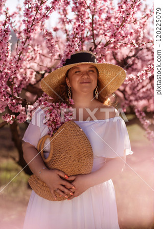 Woman blooming peach orchard. Against the backdrop of a picturesque peach orchard, a woman in a long white dress and hat enjoys a peaceful walk in the park, surrounded by the beauty of nature. 120225079