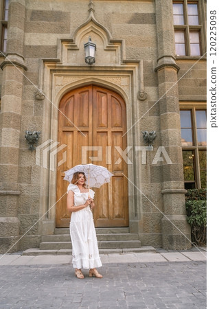 Woman Dress Umbrella Castle Entrance - A woman in a white dress holds an umbrella standing in front of the entrance to a castle. 120225098