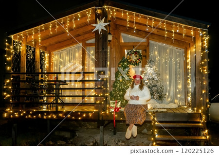Woman Christmas Lights Cabin - A woman wearing a Christmas hat stands outside a cabin decorated with Christmas lights at night. 120225126