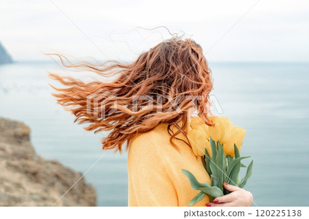 Portrait of a happy woman with hair flying in the wind against the backdrop of mountains and sea. Holding a bouquet of yellow tulips in her hands, wearing a yellow sweater Portrait of a happy woman with hair flying in the wind against the backdrop of mountains and sea. Holding a bouquet of yellow tulips in her hands, wearing a yellow sweater 120225138