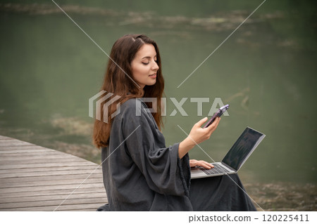 A woman is sitting on the ground with a laptop and a cell phone in her hands. She is using the laptop to browse the internet and the cell phone to communicate with someone. A woman is sitting on the ground with a laptop and a cell phone in her hands. She is using the laptop to browse the internet and the cell phone to communicate with someone. 120225411