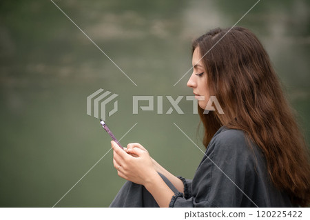 A woman is talking on her cell phone while sitting on a dock. She is wearing a black jacket and she is in a relaxed mood. A woman is talking on her cell phone while sitting on a dock. She is wearing a black jacket and she is in a relaxed mood. 120225422