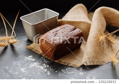 Rustic homemade rye bread loaf on burlap with wheat stalks and baking tin on dark background 120228833