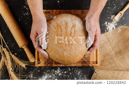 Freshly baked bread with Ukraines national emblem symbol, held on a rustic wooden board 120228842