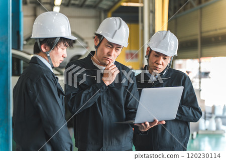 A male worker having a meeting while looking at a computer at a factory, warehouse, or logistics center (thinking, troubled, worried, investigating) 120230114
