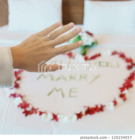 Woman hand wearing a diamond ring after say YES with message WILL YOU MARRY ME made of flower petals with bouquet on bed. Idea for marriage proposal, romantic surprise, wedding and engagement concept 120234057